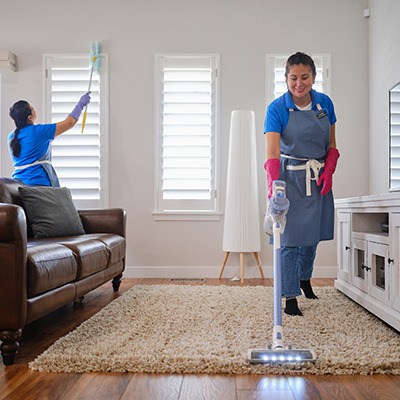 Two women wearing gloves and aprons are cleaning a living room; one is dusting shutters, and the other is vacuuming a beige rug. There is a brown leather sofa and white furniture in the bright room.