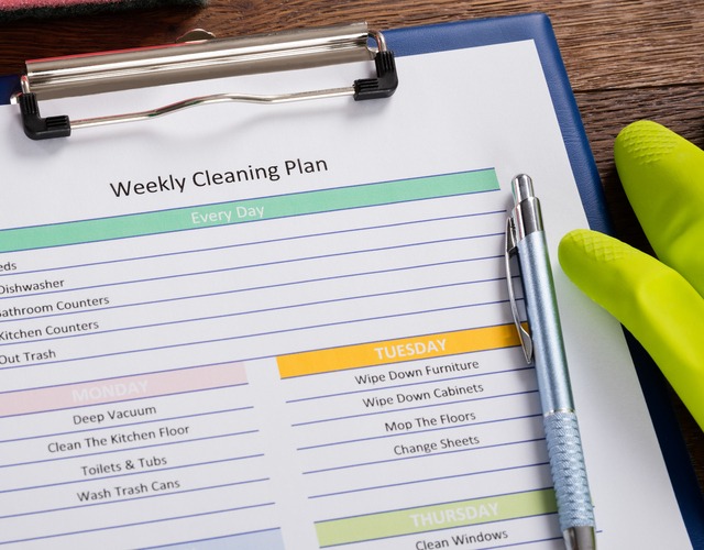 A clipboard holds a weekly cleaning plan with daily and Tuesday tasks visible. A blue pen rests on the paper, and a pair of yellow rubber cleaning gloves lies nearby on a wooden surface.
