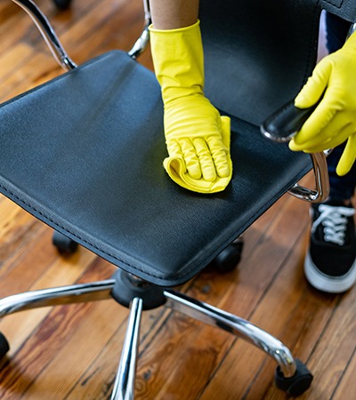 A person wearing yellow rubber gloves cleans a black office chair with a yellow cloth on a wooden floor.