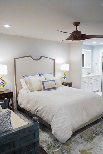 A cozy bedroom features a bed with cream bedding and multiple pillows, two bedside tables with lamps, and a ceiling fan. The space leads to a bright bathroom with a double sink vanity.