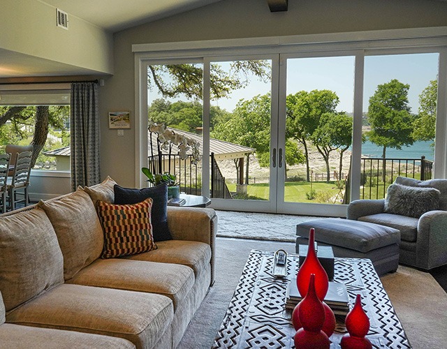 A cozy living room with a tan sectional sofa, patterned pillows, a gray armchair, and a decorative coffee table with red vases, featuring large glass doors that open to a scenic view of trees, a patio, and a lake outside.